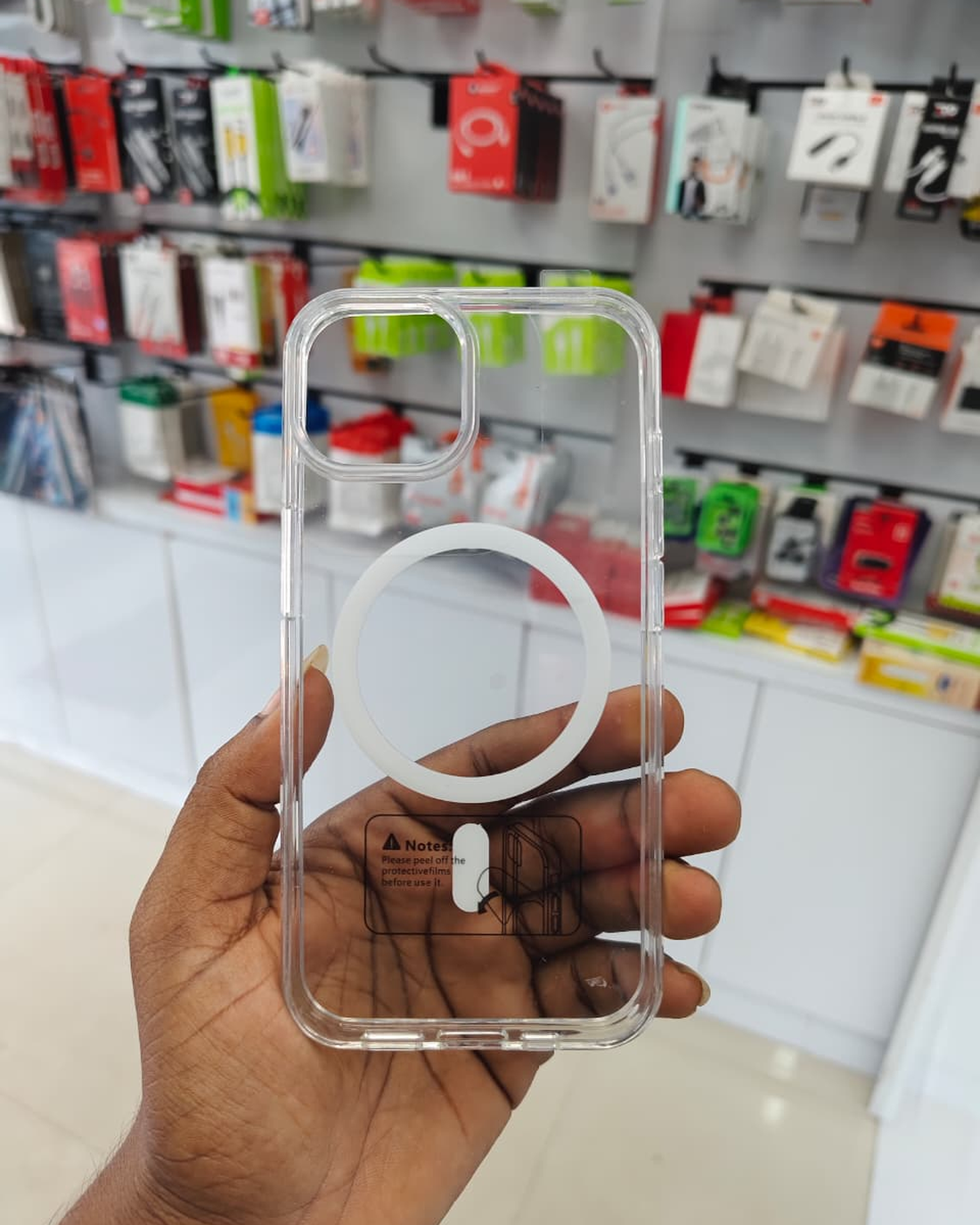 A hand holding a clear phone case with a white MagSafe ring, with a blurred electronics store background.