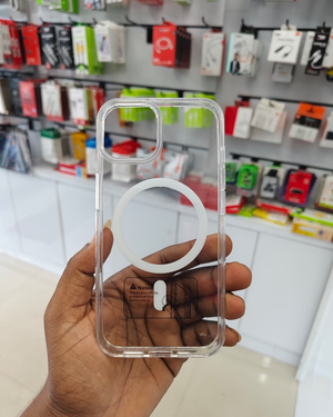 A hand holding a clear phone case with a white MagSafe ring, with a blurred electronics store background.